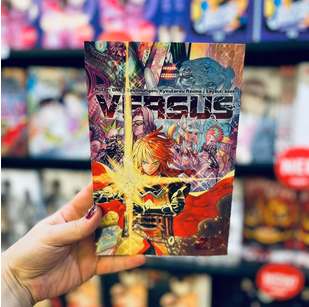 Band 2 von Versus gehalten vor einem Buchregal